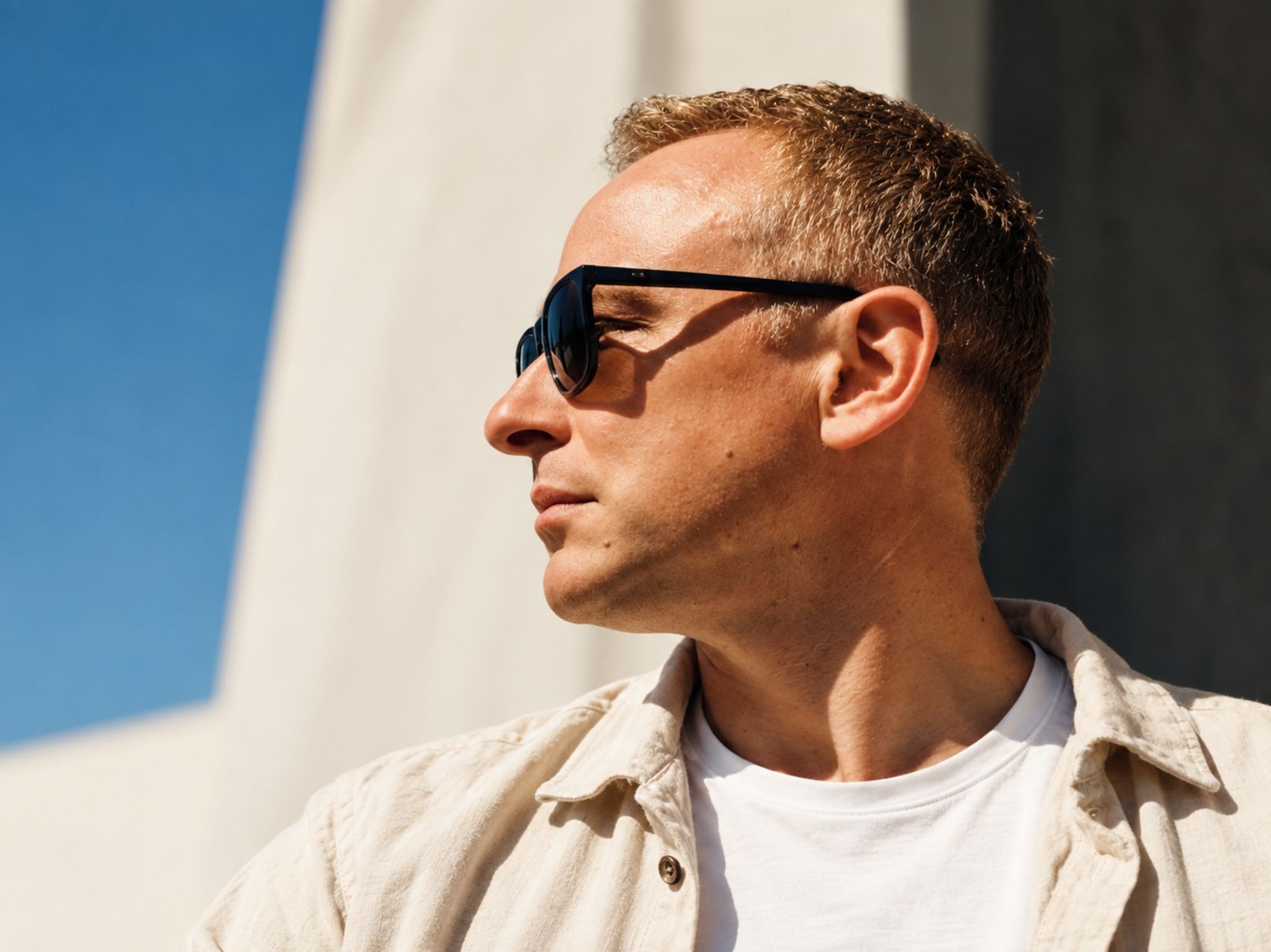 Freshwest in profile, wearing sunglasses and a linen shirt, against a sunlit pale wall and blue sky