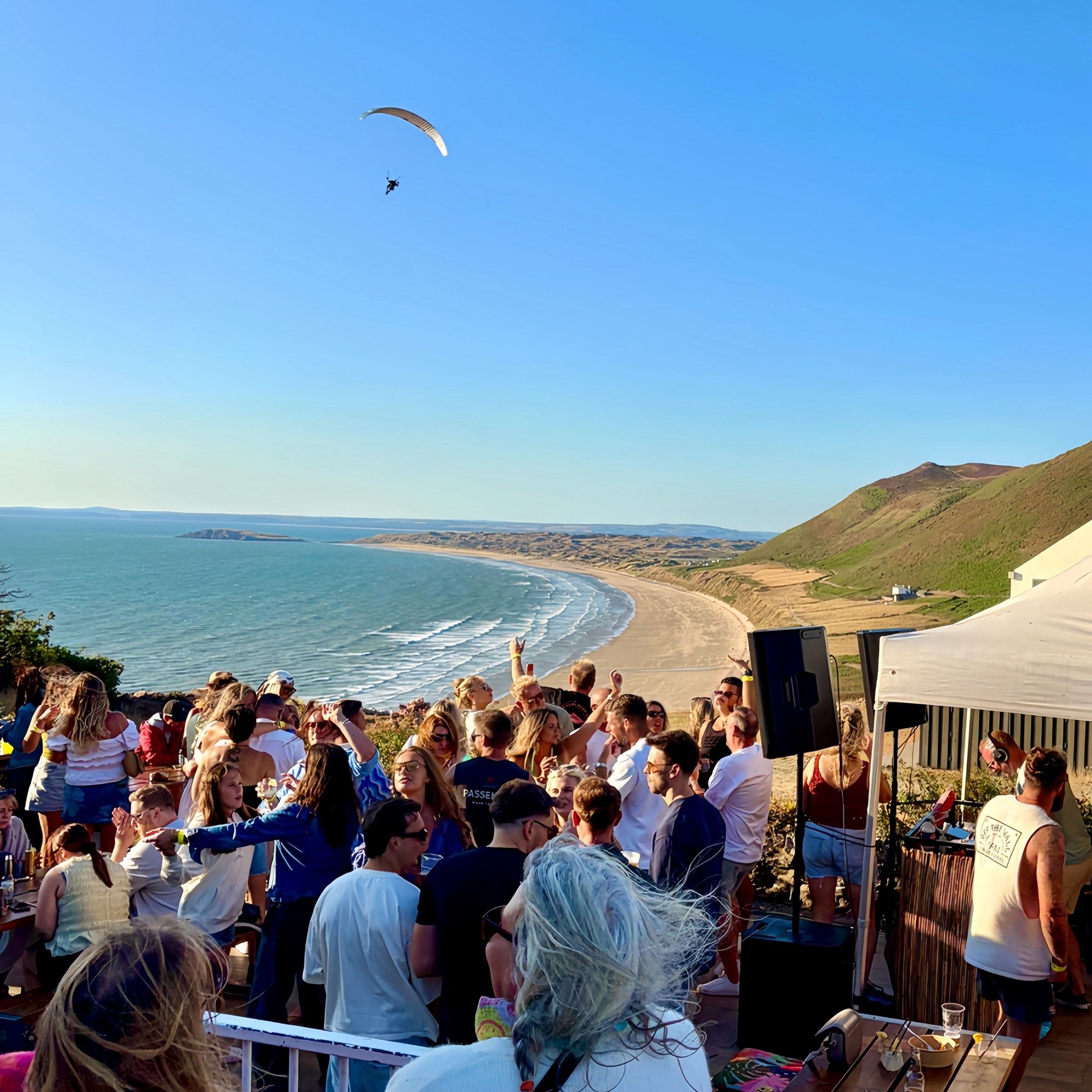 Seasunal Sessions Shoreline Soul at Rhossili, a crowd dancing on a clifftop terrace above the bay with a paraglider overhead